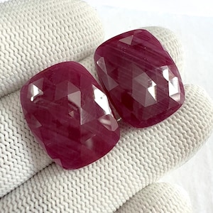 May include: Two faceted, rectangular-shaped ruby gemstones. The rubies are a deep, rich red colour with multiple facets that catch the light. The gemstones are displayed against a white, textured background.