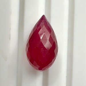 May include: A faceted, teardrop-shaped ruby gemstone. The gem is a deep, rich red colour with multiple facets that catch the light. The gemstone is set against a white background.