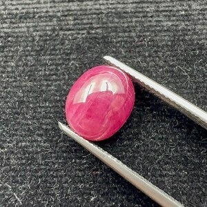 May include: A polished, oval-shaped ruby gemstone, held by silver tweezers. The ruby has a deep pinkish-red color and a smooth, reflective surface. The background is a dark, textured fabric.