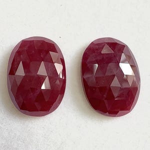 May include: Two oval-shaped, faceted rubies. The gemstones are a deep red colour, with multiple facets that reflect light. The rubies are set against a plain, light background.