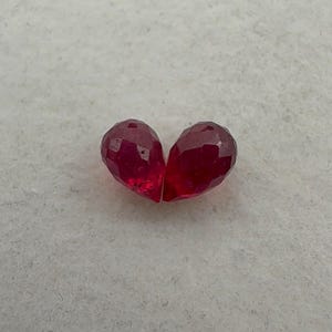 May include: Two faceted, teardrop-shaped, deep red gemstones. The gems are displayed against a neutral, textured background. The facets catch the light, creating a sparkling effect. These gems could be used for jewelry making.