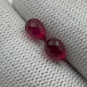 Peut inclure: Deux pierres précieuses rouges foncées, taillées en forme de goutte d'eau. Les gemmes sont présentées sur un tissu blanc texturé. Les facettes captent la lumière, créant un effet scintillant. Ces gemmes pourraient être utilisées pour la joaillerie.