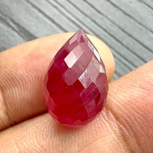 May include: A faceted, teardrop-shaped ruby gemstone. The gem is a deep, saturated red color with multiple facets that catch the light. The gemstone is held in a hand, showcasing its size and shape. The cut of the stone is designed to maximize brilliance.