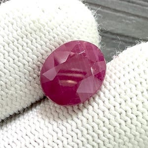 May include: A faceted, oval ruby gemstone. The gem is a deep red colour with multiple facets that reflect light. The ruby is set against a white, textured background.