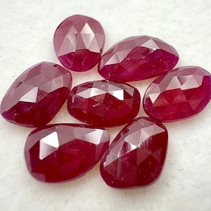 Puede incluir: Siete gemas de rubí rojo oscuro facetadas están dispuestas sobre una superficie blanca. Las gemas tienen varias formas, incluyendo ovaladas y de pera, y reflejan la luz, mostrando su talla y color. Estos rubíes son adecuados para la fabricación de joyas.