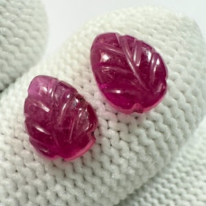Peut inclure: Deux pierres précieuses en rubis sculptées en forme de feuilles. Les pierres précieuses sont d'un rose profond et présentent des veines détaillées. Les gemmes sont exposées sur une surface blanche et texturée.