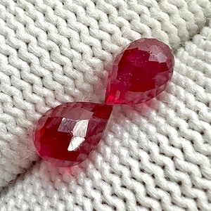 May include: Two faceted, teardrop-shaped, deep red gemstones. The gems are displayed on a white, textured, knitted fabric. The gems have multiple facets that reflect light, creating a sparkling effect. The overall theme is jewelry and gemstones.