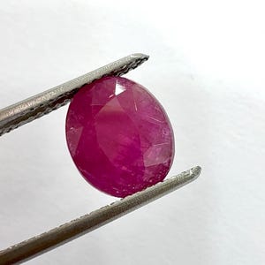 Puede incluir: Una gema de rubí ovalada y facetada, sujeta con pinzas plateadas. El rubí es de un color rojo intenso y saturado, con inclusiones internas visibles y una superficie pulida. Las pinzas están enfocadas sobre un fondo blanco.