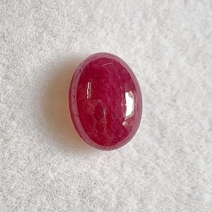 May include: A polished, oval ruby gemstone. The gem is a deep red colour with a glossy surface. The stone is set against a white, textured background.