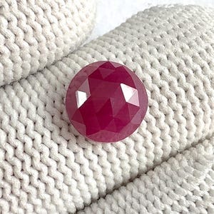 Puede incluir: Una gema facetada, redonda, de color rojo intenso. La gema tiene un corte brillante con muchas superficies reflectantes. La piedra se encuentra sobre un fondo de punto blanco roto texturizado, resaltando su color y claridad. Adecuado para joyería.