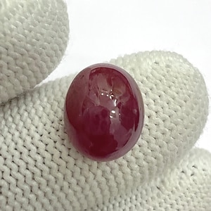May include: A polished, oval ruby gemstone. The gem is a deep red colour with a glossy surface. The ruby is held against a white, textured background.