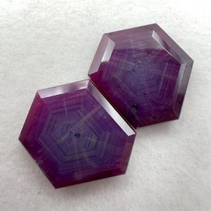 Puede incluir: Dos piedras preciosas hexagonales de color magenta intenso que presentan distintos patrones geométricos concéntricos en su estructura. Las piedras tienen superficies planas y pulidas con bandas internas visibles que crean un efecto.