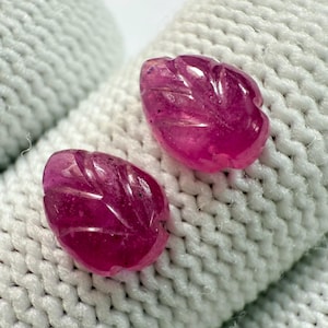 May include: Two carved, fuchsia-coloured gemstones, each shaped like a leaf, rest on a white, textured surface. The gemstones have detailed veining and a polished appearance, showcasing their vibrant colour and intricate design.
