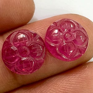 Może przedstawiać: Dwa okrągłe, wypolerowane, rubinowo-czerwone kamienie szlachetne. Każdy kamień szlachetny ma rzeźbiony kwiatowy wzór z centralnym okręgiem i otaczającymi płatkami. Kamienie szlachetne są trzymane między palcami, ukazując ich żywy kolor i skomplikowane detale.