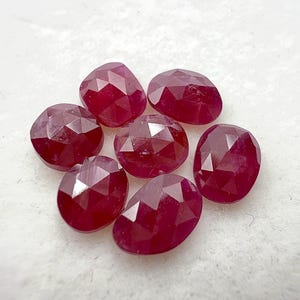 May include: Seven faceted, oval-shaped ruby gemstones. The rubies are a deep, rich red color and have multiple facets that catch the light. The gemstones are arranged in a circular pattern against a white background.
