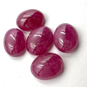 Könnte beinhalten: Fünf ovale, polierte Rubin-Edelsteine. Die Rubine haben eine tiefe, satte rote Farbe mit glänzender Oberfläche. Die Edelsteine sind auf weißem Hintergrund angeordnet, um ihre Farbe und Form hervorzuheben.