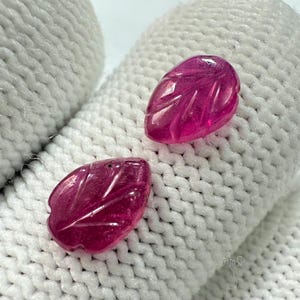 May include: Two magenta-coloured, leaf-shaped gemstones with carved veining. The polished stones are displayed against a white, textured background. These gemstones are suitable for jewellery making or collecting.