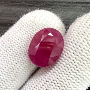 Peut inclure: Une pierre précieuse de rubis ovale et facettée. Le rubis est d'une couleur rouge profond et riche, avec de multiples facettes qui réfléchissent la lumière. La pierre est tenue sur un fond blanc texturé.