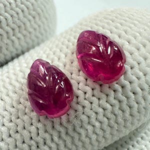 Puede incluir: Dos gemas de rubí talladas, cada una con forma de hoja, se exhiben sobre una superficie blanca texturizada. Las gemas son de un color rojo-rosa intenso y vibrante, con intrincados detalles tallados.