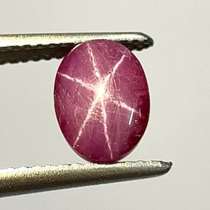 Puede incluir: Una gema de rubí pulida, de forma ovalada, sujeta con unas pinzas. La gema es de color rojo intenso con un patrón de estrella de seis rayos en su superficie. El efecto de estrella se crea por la reflexión de la luz.