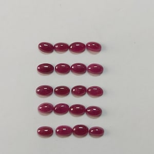 Puede incluir: Conjunto de dieciséis gemas de rubí ovaladas y pulidas. Las piedras son de un color rojo intenso y están dispuestas en filas sobre una superficie blanca. Las gemas varían ligeramente en tamaño, mostrando su belleza natural.
