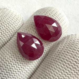 May include: Two faceted, teardrop-shaped rubies. The gems are a deep, rich red colour and have multiple facets that catch the light. The rubies are displayed on a white, textured surface.