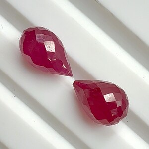 May include: Two faceted, teardrop-shaped gemstones in a deep red hue. The gems have multiple facets that catch the light, creating a sparkling effect. The stones are displayed on a white surface.