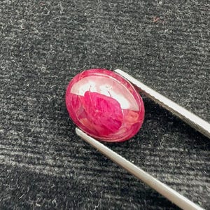 May include: A cabochon-cut ruby gemstone, oval in shape. The deep red gem is held by silver tweezers, set against a dark, textured backdrop. The ruby displays internal inclusions and a polished surface that reflects light.
