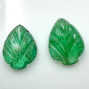 May include: Two carved emerald green gemstone leaf pendants. The leaves have detailed veining and a polished finish. The gemstones are set against a white background.