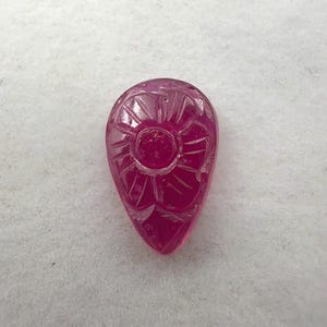 May include: A teardrop-shaped, carved ruby gemstone. The gemstone features a central floral design with radiating lines. The ruby is a deep, translucent red color and is set against a white background.