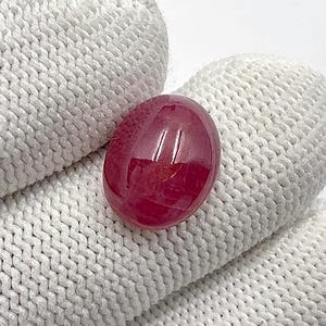 May include: A polished, oval-shaped, deep red gemstone. The gem has a smooth, reflective surface and appears to be set against a white, textured background. The stone's colour is a rich, saturated red, with subtle variations in tone.