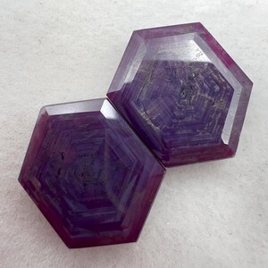 Op de afbeelding: Twee gefacetteerde zeshoekige edelstenen met een dieppaars en magenta kleurenpalet. Elke steen vertoont een duidelijk geometrisch patroon van concentrische zeshoekige banden die vanuit het midden uitstralen, met een gladde, gepolijste afwerking.