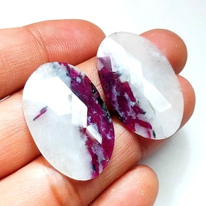 May include: Two oval-shaped, white gemstones with red and black inclusions. The gemstones are smooth and polished.