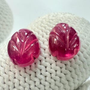 Könnte beinhalten: Zwei geschnitzte, oval geformte, rubinrote Edelsteine. Jeder Edelstein weist ein blattartiges Design mit detaillierten Adern auf. Die Edelsteine werden auf einer weißen, strukturierten Oberfläche präsentiert.