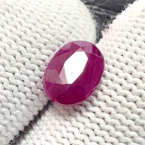 Puede incluir: Una gema de rubí ovalada y facetada. La gema es de un color rojo intenso y rico con múltiples facetas que reflejan la luz. El rubí está sobre un fondo blanco texturizado.