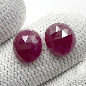Pode incluir: Duas pedras preciosas de rubi facetadas em forma oval. Os rubis são de uma cor vermelha profunda e rica e têm múltiplas facetas que capturam a luz. São exibidos em uma superfície branca e texturizada.