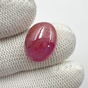 Op de afbeelding: Een gepolijste, ovaalvormige edelsteen met een levendige roze en rode tint. De edelsteen is glad en reflecterend, met een glanzend oppervlak. De steen wordt vastgehouden in een witte gebreide handschoen, die de kleur en vorm benadrukt.
