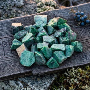 May include: A pile of rough-cut green and red ruby zoisite stones on a weathered wooden surface. The stones are irregularly shaped and vary in size. The background includes a sprig of blue berries and some moss.