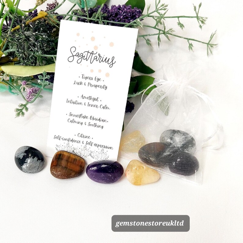 May include: A collection of polished gemstones, including Tiger's Eye, Amethyst, and Citrine, with a white card labeled "Sagittarius." The card lists properties associated with each stone. The gemstones are displayed with a small white organza bag and sprigs of greenery.