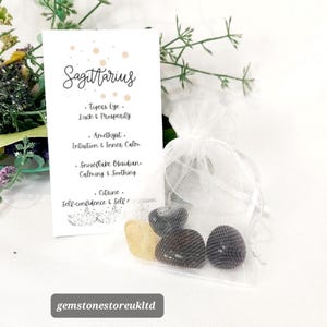 May include: A white organza bag filled with gemstones, next to a card with the word "Sagittarius" and gemstone descriptions. The gemstones are yellow, black, and purple. The card lists the gemstones and their properties.
