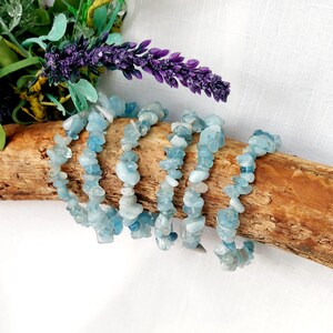 K&ouml;nnte beinhalten: Sechs Armb&auml;nder aus Aquamarin-Chip-Perlen werden auf einem St&uuml;ck Treibholz pr&auml;sentiert. Die Armb&auml;nder bestehen aus kleinen, unregelm&auml;&szlig;igen, hellblauen und wei&szlig;en Steinen. Der Hintergrund umfasst lila Lavendel und gr&uuml;nes Laub vor einem wei&szlig;en Hintergrund.