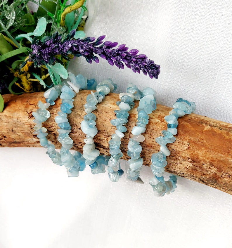 K&ouml;nnte beinhalten: Sechs Armb&auml;nder aus Aquamarin-Chip-Perlen werden auf einem St&uuml;ck Treibholz pr&auml;sentiert. Die Armb&auml;nder bestehen aus kleinen, unregelm&auml;&szlig;igen, hellblauen und wei&szlig;en Steinen. Der Hintergrund umfasst lila Lavendel und gr&uuml;nes Laub vor einem wei&szlig;en Hintergrund.