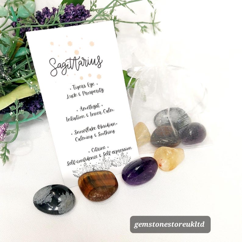 May include: A collection of polished gemstones, including Tiger's Eye, Amethyst, and Citrine, arranged with a card labeled "Sagittarius." The card lists the properties of each stone. A small, sheer bag contains additional stones. The background includes greenery.