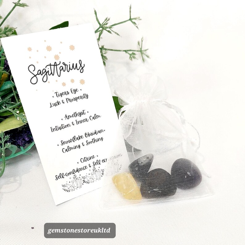 May include: A white organza bag filled with gemstones, next to a white card with the word "Sagittarius" and a list of associated stones and properties. The gemstones include tiger's eye, amethyst, snowflake obsidian, and citrine.