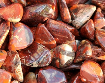 Op de afbeelding: Een close-up van een verzameling rode jaspis-tumbled stones. De stenen zijn glad en gepolijst, met een verscheidenheid aan rode, bruine en witte patronen.
