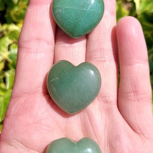 Pu&ograve; includere: Tre pietre di avventurina verde a forma di cuore sono esposte in una mano. Le pietre hanno una superficie liscia e lucida e variano leggermente nella tonalit&agrave;. Lo sfondo &egrave; sfocato, con fogliame verde visibile.