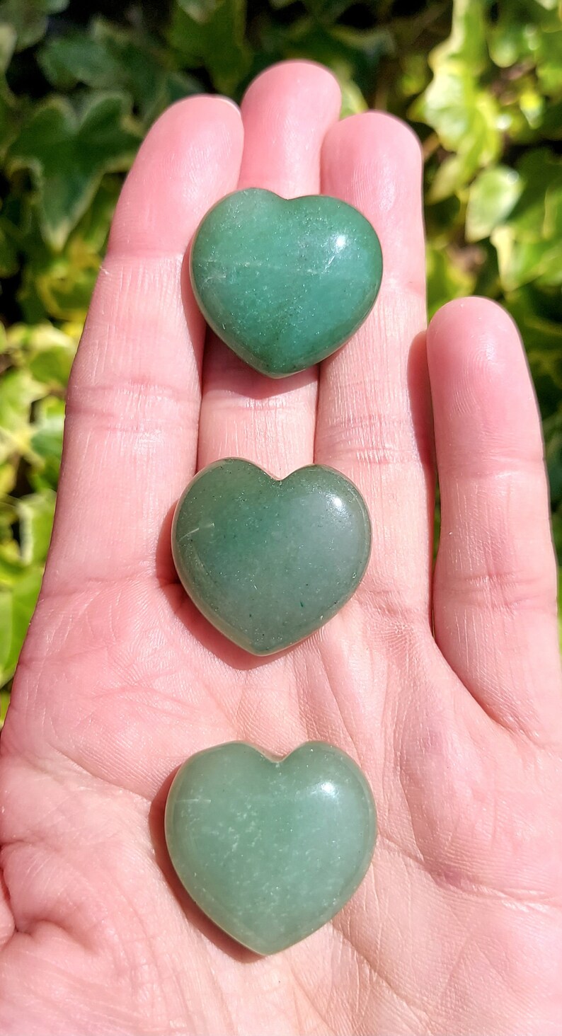 Pu&ograve; includere: Tre pietre di avventurina verde a forma di cuore sono esposte in una mano. Le pietre hanno una superficie liscia e lucida e variano leggermente nella tonalit&agrave;. Lo sfondo &egrave; sfocato, con fogliame verde visibile.