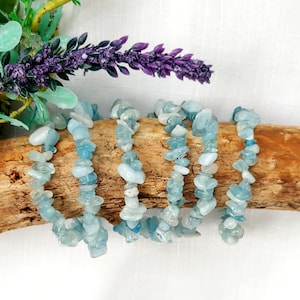 K&ouml;nnte beinhalten: Mehrere Armb&auml;nder aus Aquamarin-Chip-Perlen werden auf einem St&uuml;ck Treibholz pr&auml;sentiert. Die Armb&auml;nder sind mit hellblauen und wei&szlig;en Steinen besetzt. Im Hintergrund befinden sich lila Lavendel und gr&uuml;ne Bl&auml;tter.