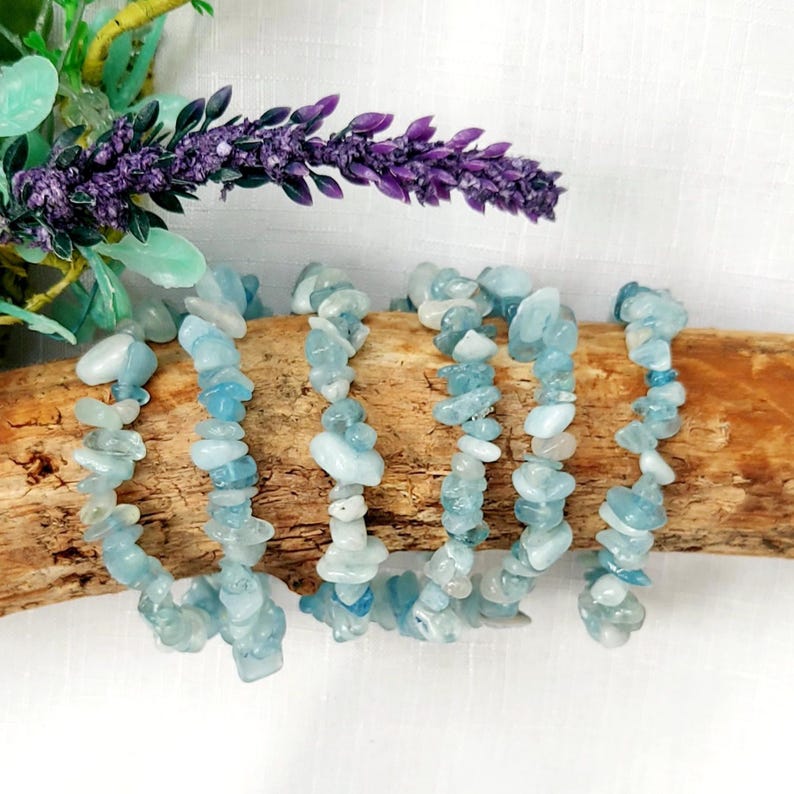 K&ouml;nnte beinhalten: Mehrere Armb&auml;nder aus Aquamarin-Chip-Perlen werden auf einem St&uuml;ck Treibholz pr&auml;sentiert. Die Armb&auml;nder sind mit hellblauen und wei&szlig;en Steinen besetzt. Im Hintergrund befinden sich lila Lavendel und gr&uuml;ne Bl&auml;tter.