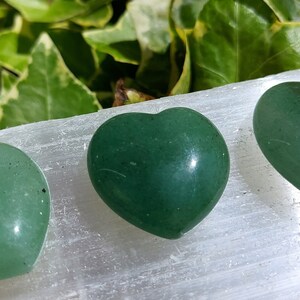 Pu&ograve; includere: Tre pietre a forma di cuore verde lucido. Le pietre sono disposte su una superficie bianca, con fogliame verde sullo sfondo. I cuori hanno una finitura liscia e lucida e variano leggermente nella forma.